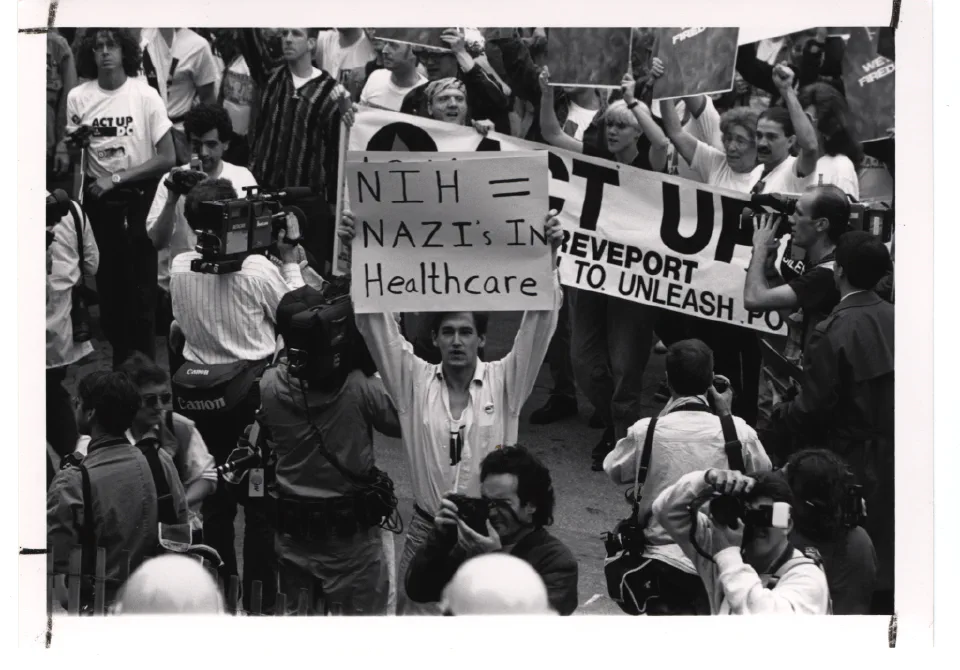 मई 1990 में अमेरिकी राष्ट्रीय स्वास्थ्य संस्थान (NIH) के समक्ष प्रदर्शन करते ACT UP आंदोलन के कार्यकर्ता। फोटो: [NIH History Office / Wikimedia Commons](https://commons.wikimedia.org/wiki/File:ACT_UP_Activists_at_the_National_Institutes_of_Health_(14359361475).jpg) (पब्लिक डोमेन)